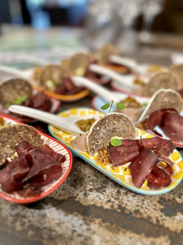 Häppchen mit Brot, Fleisch und Senf werden auf bunten Löffeln serviert.