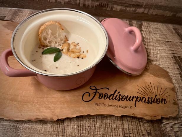 Eine cremige Suppe mit Brot und Salbei in einem rosa Topf auf einem Holzbrett.