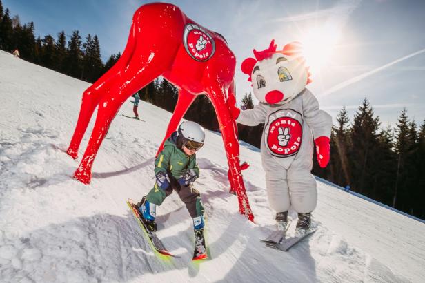 Ein Kind fährt Ski neben einer roten Hirschstatue und dem Maskottchen von Ben Monte Popolo.