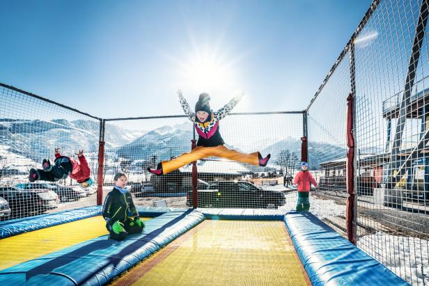 Kinder springen auf einem Trampolin vor einer verschneiten Bergkulisse.