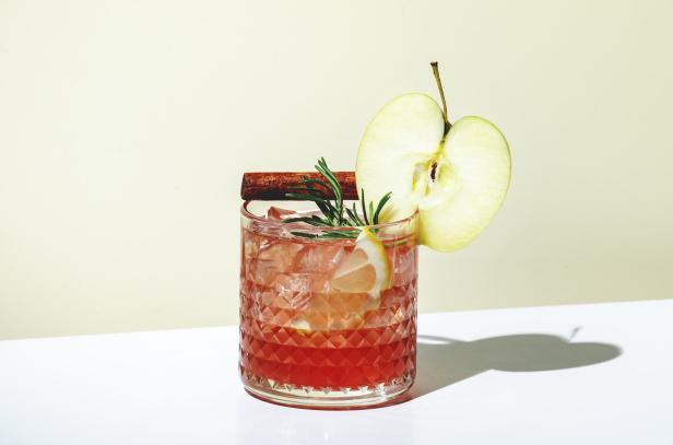 Ein Cocktail mit Apfel, Zimtstange und Rosmarin auf Eis in einem verzierten Glas.