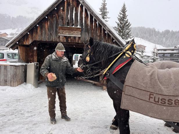 Ein Mann füttert ein schwarzes Pferd im Schnee vor einem Holzgebäude.