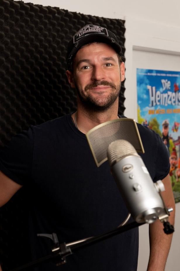 Ein Mann mit Bart und Kappe steht vor einem Mikrofon, im Hintergrund ein Poster von „Die Heinzels“.