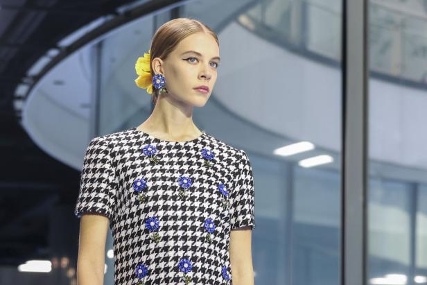 Eine Frau mit blonden Haaren trägt ein Hahnentrittkleid mit blauen Blumen und eine gelbe Blume im Haar.