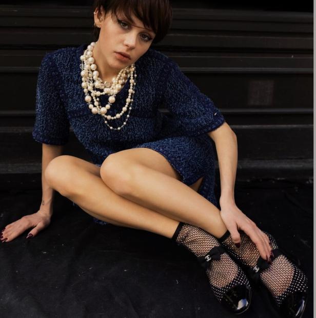 Eine junge Frau mit kurzem Haar trägt ein blaues Kleid und eine Perlenkette.