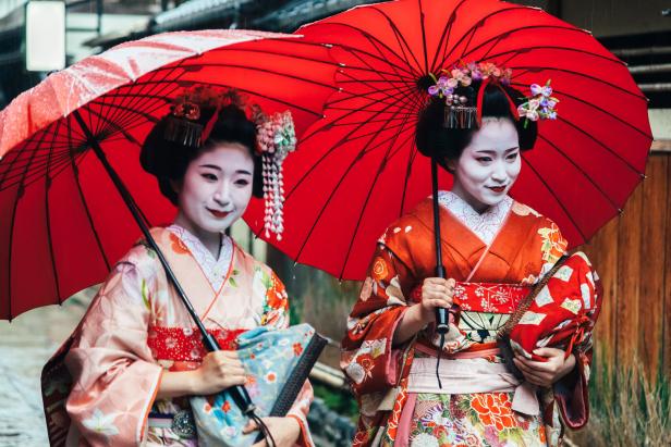 Zwei Geishas mit traditioneller Kleidung und roten Schirmen im Regen.