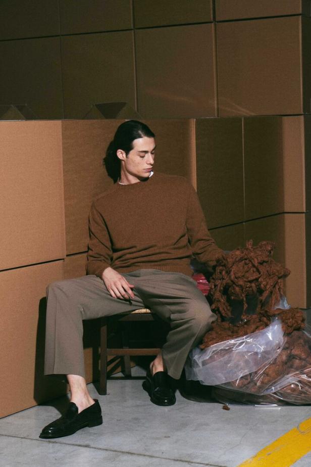 Ein Mann mit braunem Pullover und grauer Hose sitzt auf einem Stuhl neben einem Sack mit braunem Material.