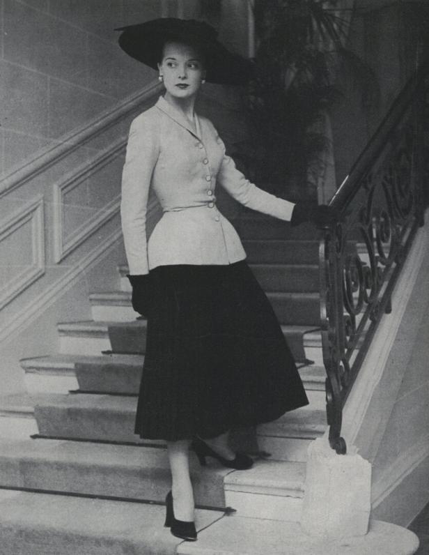 Eine Frau in einem eleganten Kostüm und einem großen Hut auf einer Treppe.