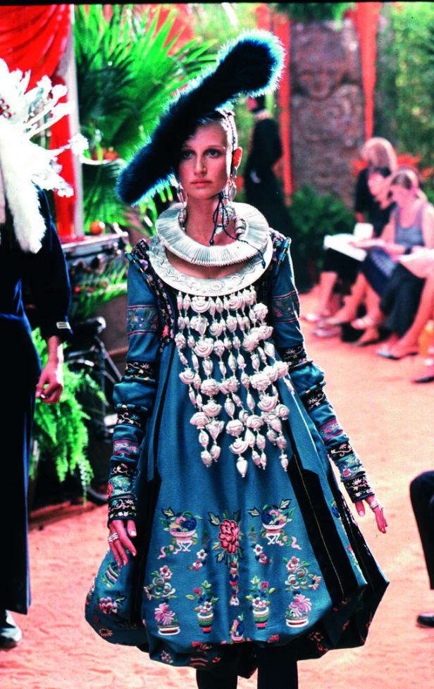 Eine Frau in einem blauen Kleid mit Blumenmuster und einem großen Federschmuck auf einer Modenschau.