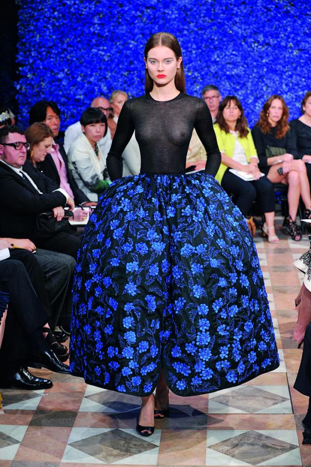 Ein Model präsentiert ein schwarzes Kleid mit blauen Blumen auf einem Laufsteg.