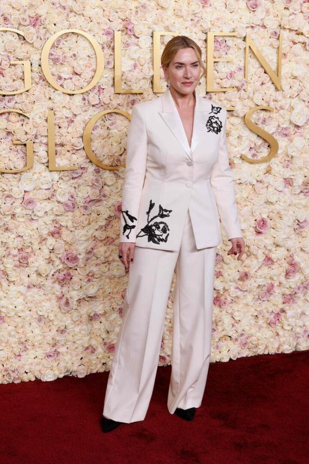 Kate Winslet in einem weißen Hosenanzug auf dem roten Teppich vor einer Blumenwand.