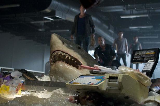 Ein Hai durchbricht eine Kasse in einem überschwemmten Supermarkt, während Menschen im Hintergrund stehen.