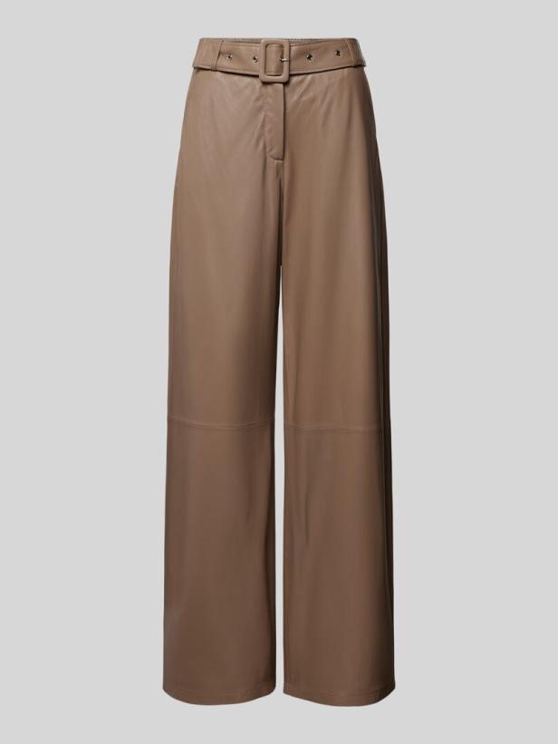 Eine beigefarbene, weite Lederhose mit Gürtel vor einem grauen Hintergrund.