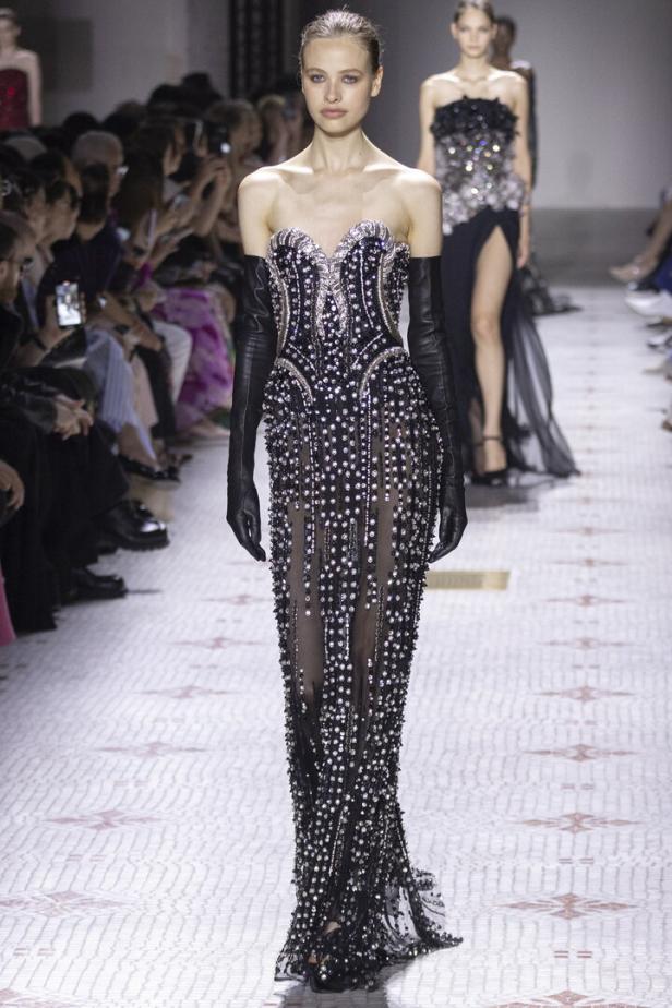 Ein Model präsentiert ein trägerloses, schwarzes Abendkleid mit silbernen Verzierungen und langen Handschuhen auf dem Laufsteg.