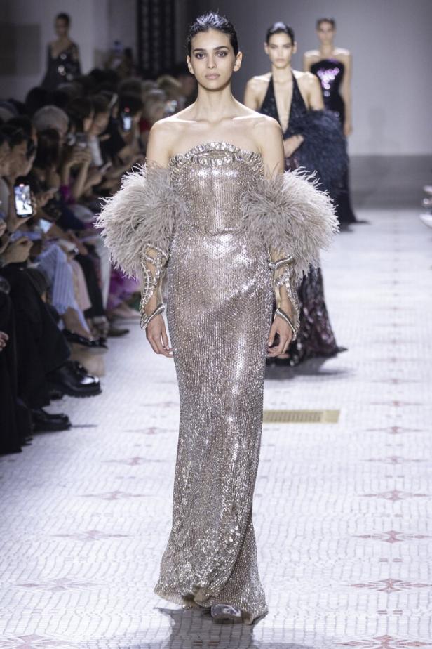 Ein Model präsentiert ein trägerloses, silbernes Paillettenkleid mit Federdetails auf dem Laufsteg.