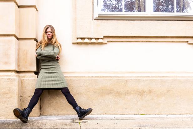 Zoë posiert in einem grünen Kleid und schwarzen Stiefeln an einer Wand.