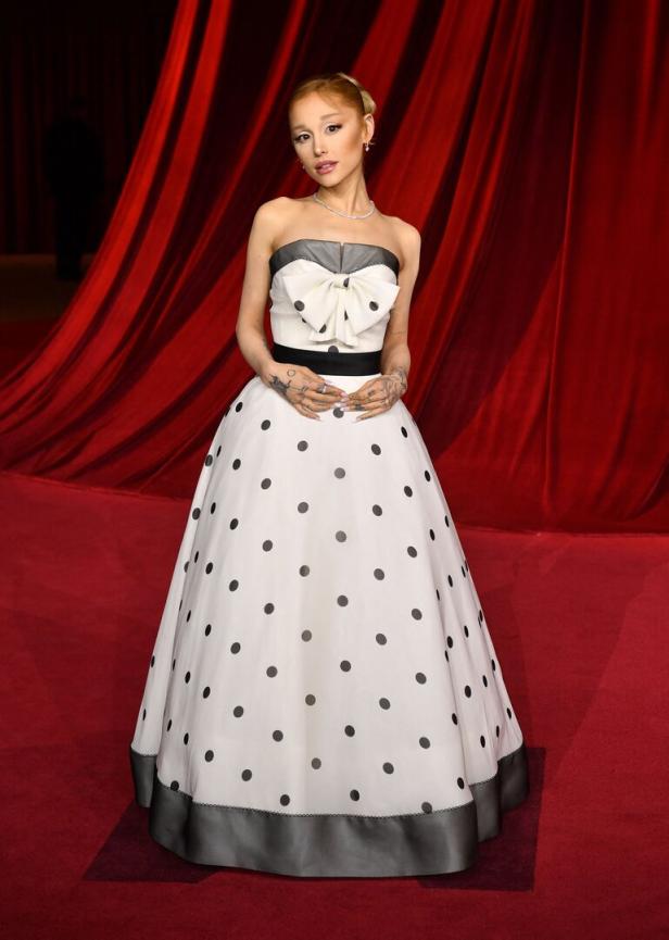 Ariana Grande in einem weißen, gepunkteten Kleid mit Schleife vor einem roten Vorhang.