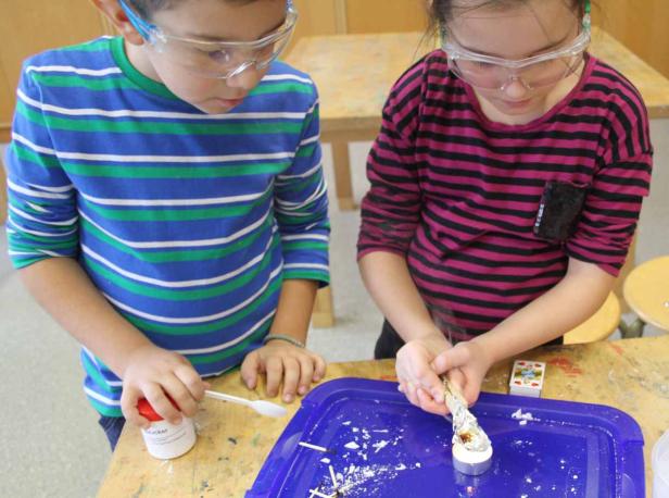 Zwei Kinder mit Schutzbrillen experimentieren mit einer Kerze und Chemikalien.
