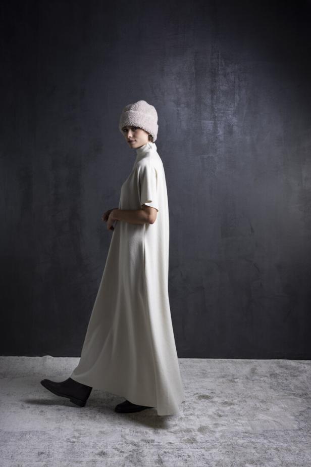 Eine Frau in einem langen, weißen Kleid und einer Fellmütze geht vor einer dunklen Wand.