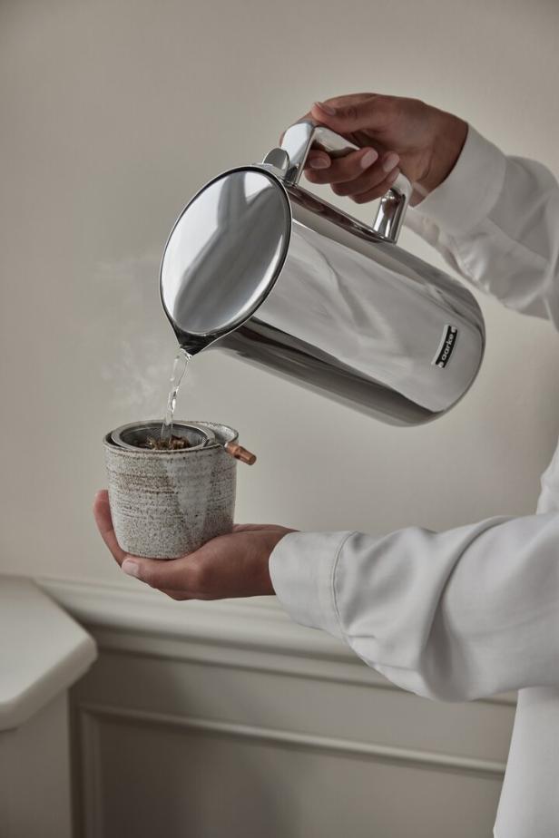 Eine Person gießt Wasser aus einer silbernen Kanne in eine Tasse mit einem Teesieb.
