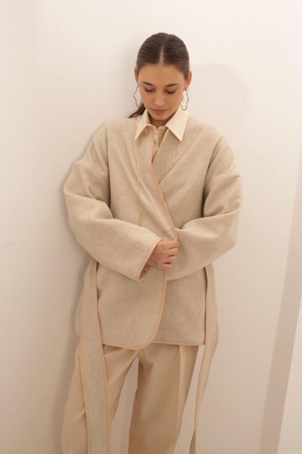 Eine Frau in einem beigen Outfit mit übergroßem Mantel und goldenen Ohrringen steht vor einer Wand.
