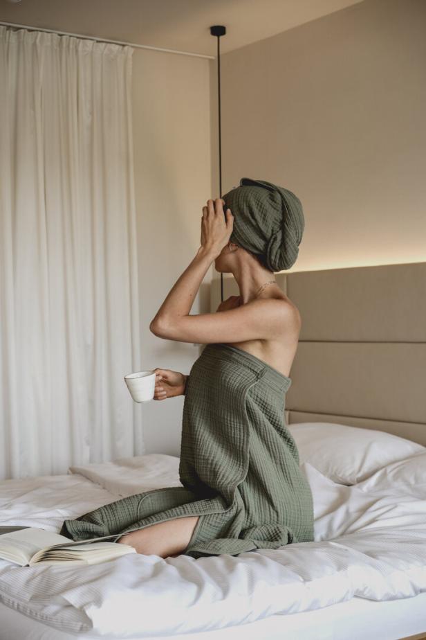 Eine Frau sitzt in ein Handtuch gehüllt auf einem Bett und hält eine Tasse in der Hand.
