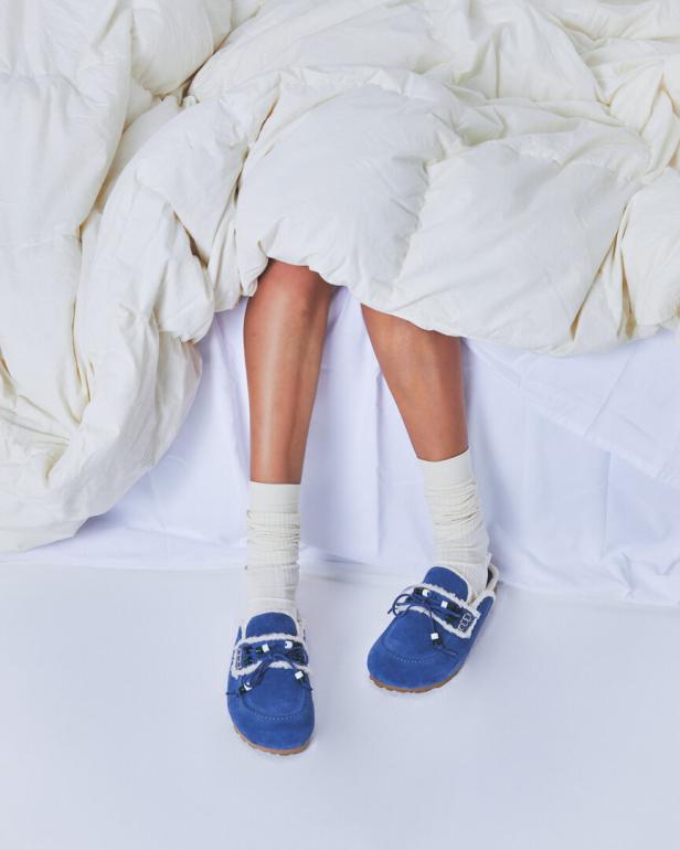 Eine Person liegt mit Socken und blauen Hausschuhen im Bett unter einer weißen Decke.