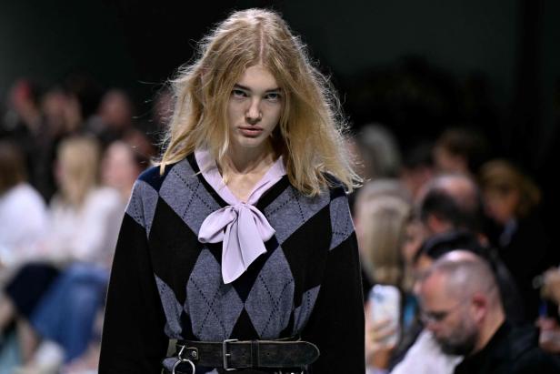 Eine blonde Frau mit einem Argyle-Pullover und einer Schleife steht vor einer Zuschauermenge.