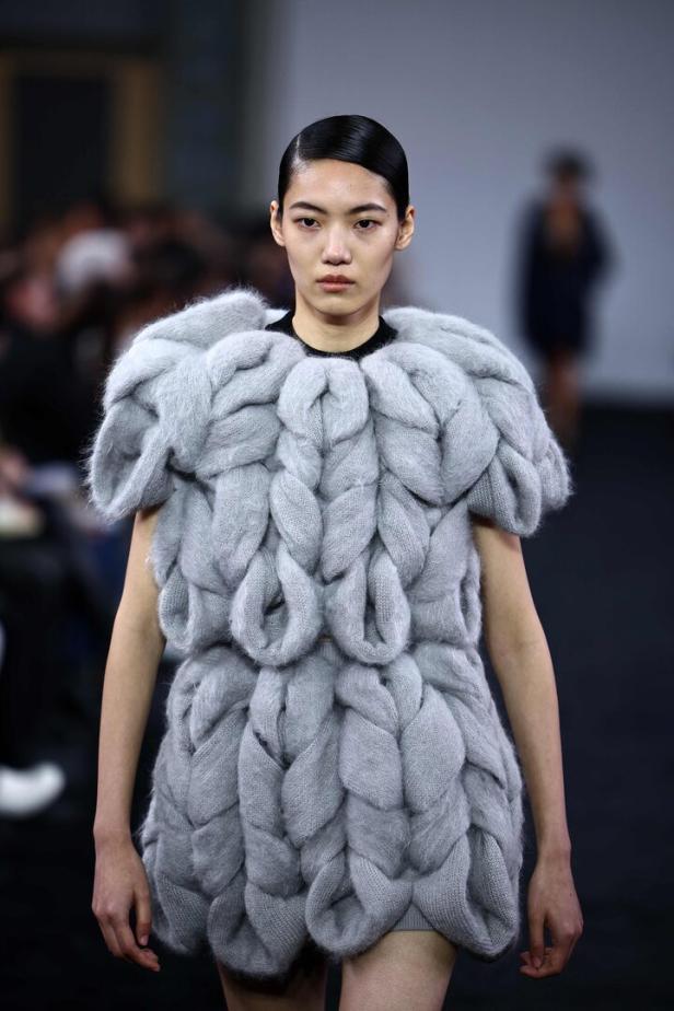 Eine Frau präsentiert ein Kleid aus grobem, grauem Strick auf einem Laufsteg.