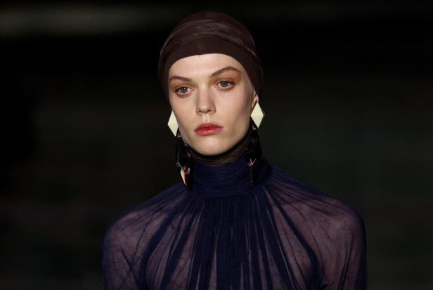 Ein Model mit braunem Turban und transparentem, hochgeschlossenem Oberteil trägt große, geometrische Ohrringe.