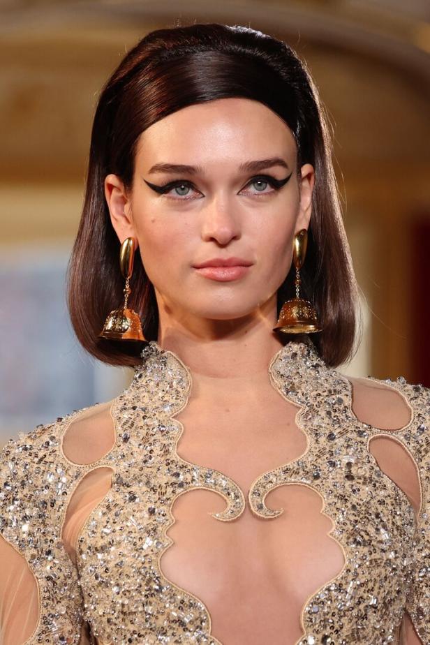 Eine Frau mit auffälligem Eyeliner und goldenen Ohrringen in Glockenform trägt ein verziertes Kleid.