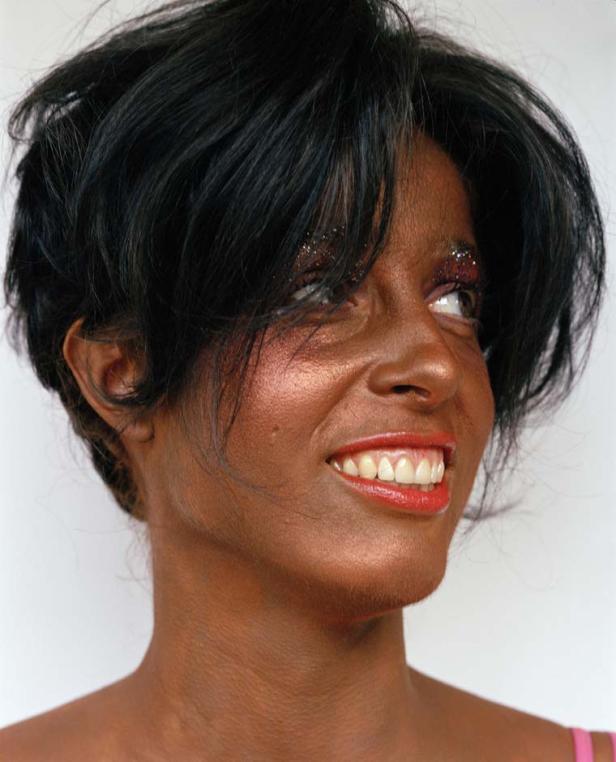 Eine Frau mit dunkler Haut, glitzerndem Make-up und kurzem, schwarzem Haar lächelt.