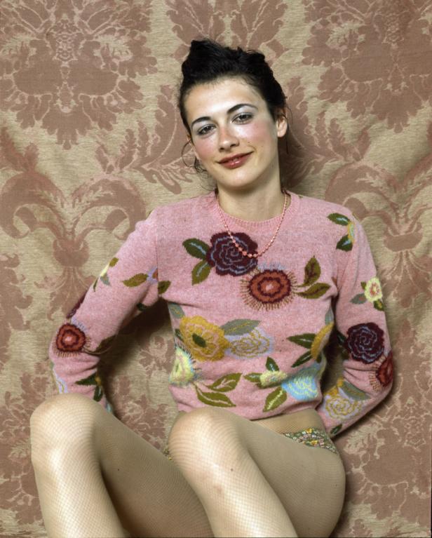 Eine junge Frau mit rosa Pullover und Blumenmuster posiert vor einem gemusterten Hintergrund.