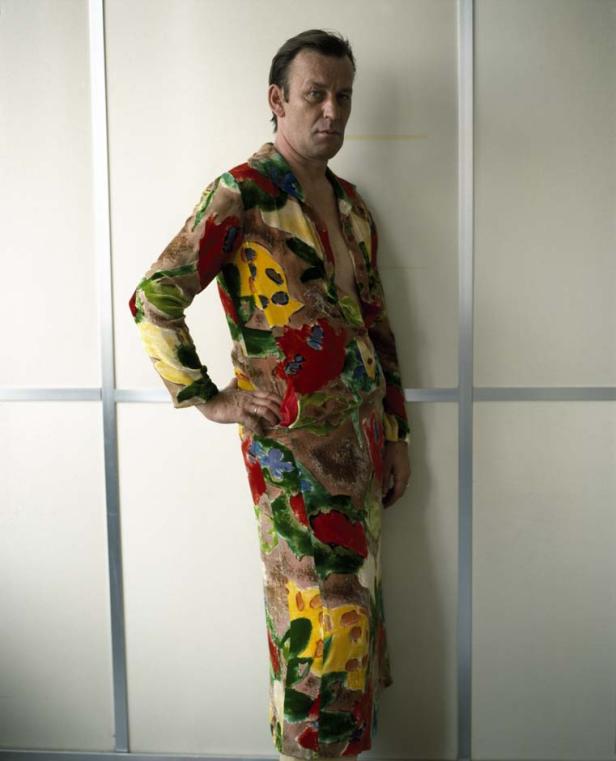 Ein Mann posiert in einem farbenfrohen, gemusterten Kleid vor einer weißen Wand.