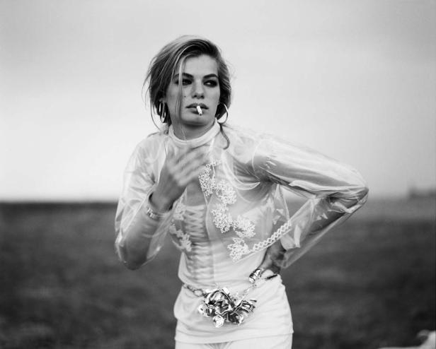 Eine Frau mit Zigarette, gekleidet in eine transparente Jacke, steht im Freien.