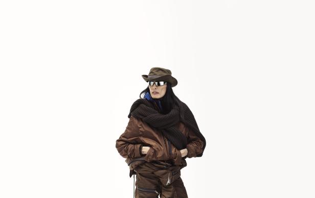 Eine Frau mit Hut, Sonnenbrille und brauner Jacke posiert vor einem weißen Hintergrund.