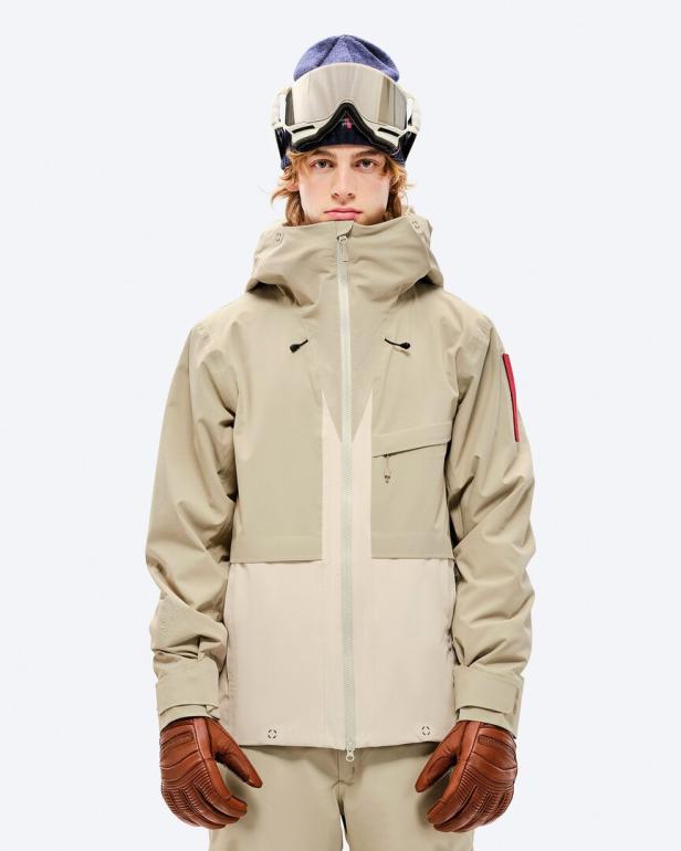 Ein Mann in Skikleidung mit Mütze, Skibrille und Handschuhen vor einem weißen Hintergrund.