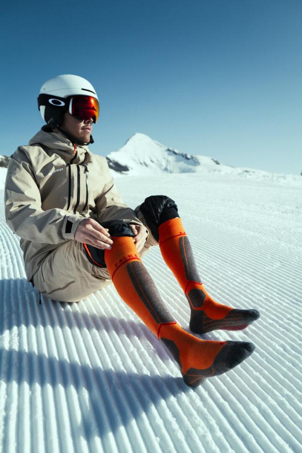 Ein Skifahrer mit Helm und Skibrille sitzt auf der Piste und trägt orangefarbene Socken.