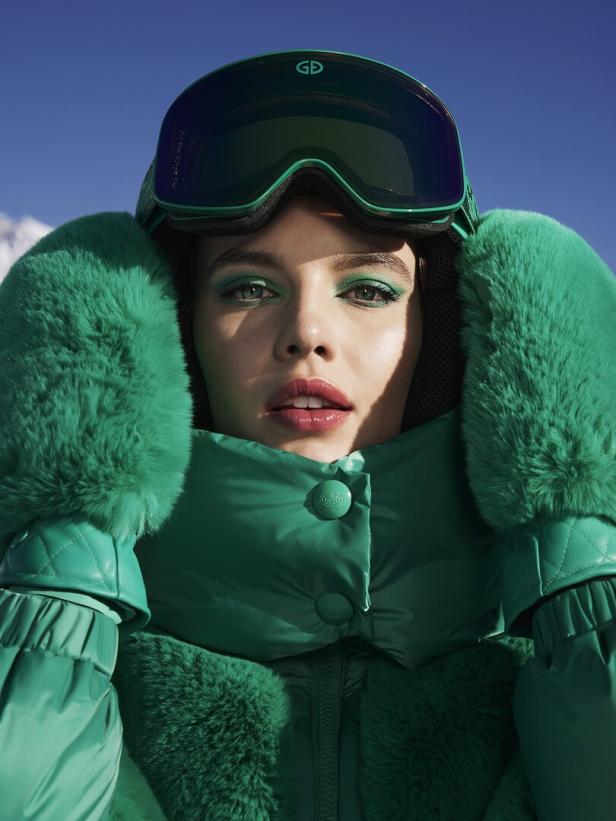 Eine Frau mit grüner Skijacke, Fäustlingen und Skibrille vor blauem Himmel.