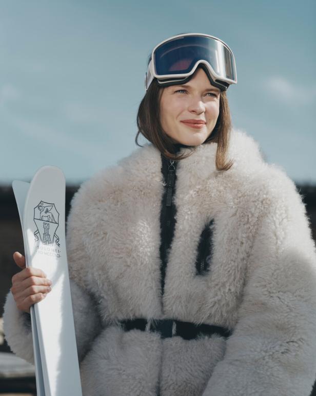 Eine Frau in flauschiger Kleidung und Skibrille hält ein Paar Ski.