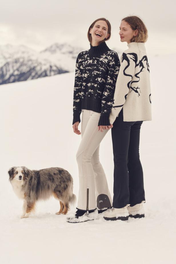 Zwei Frauen stehen mit einem Hund im Schnee, im Hintergrund sind Berge zu sehen.