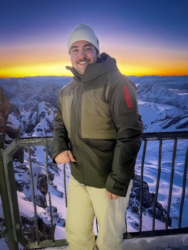 Ein lächelnder Mann in Winterkleidung vor einem Bergpanorama bei Sonnenaufgang.