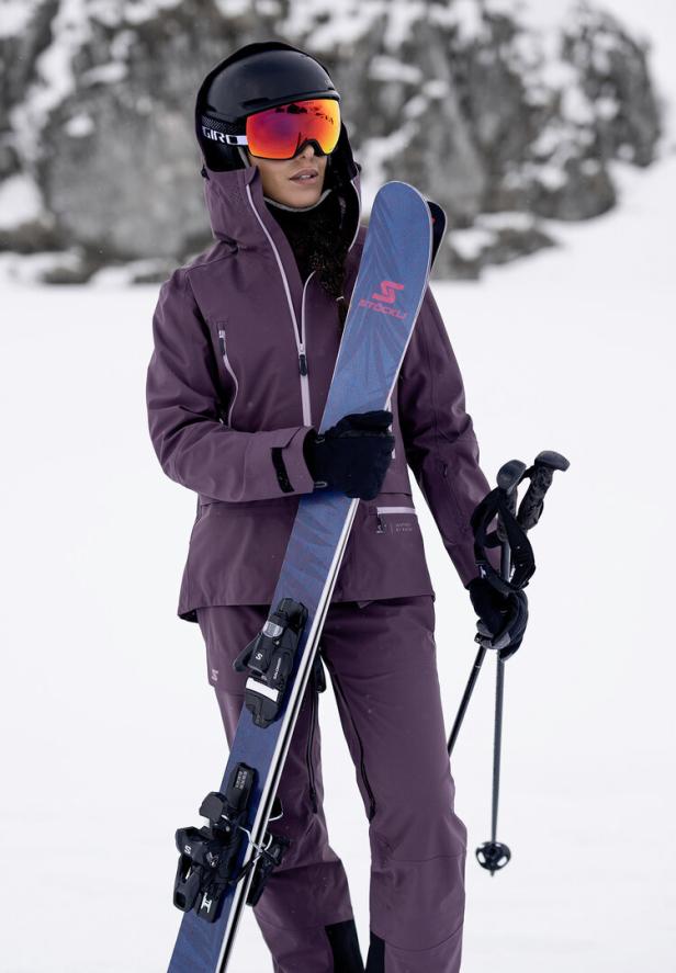 Eine Frau in Skikleidung hält Ski und Stöcke im Schnee.