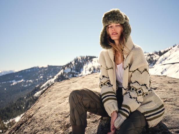 Eine Frau sitzt auf einem Felsen in den Bergen und trägt eine Pelzmütze und einen Strickjacke.