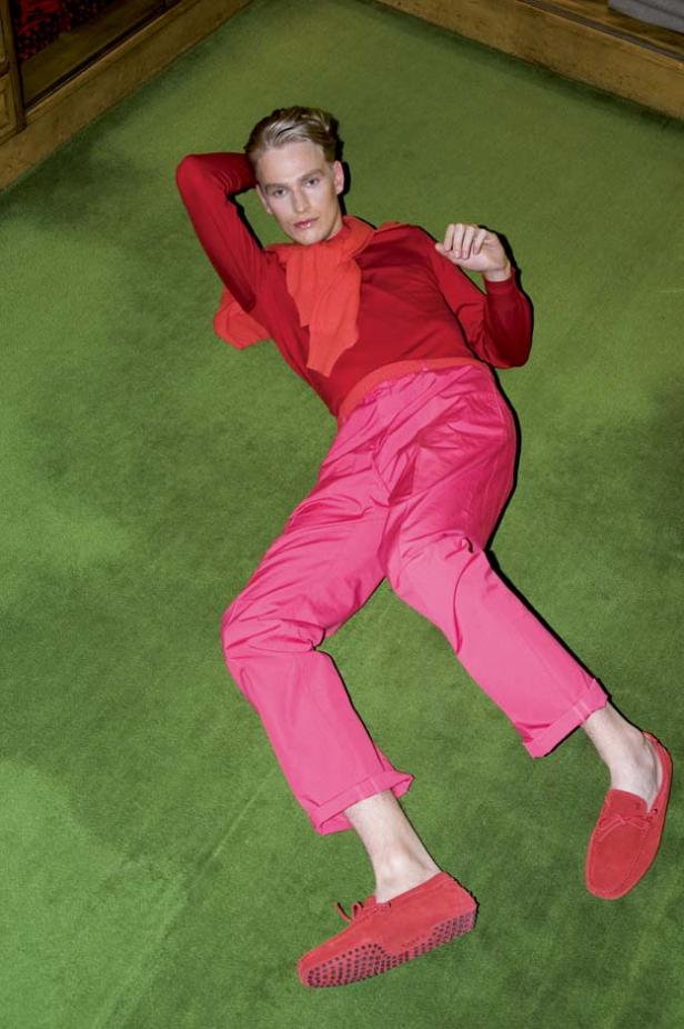 Ein Mann liegt in roter Kleidung und pinkfarbener Hose auf einem grünen Teppich.