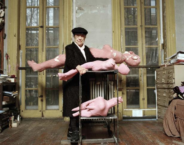 Eine ältere Frau mit einer schwarzen Baskenmütze posiert mit rosa Stoffpuppen in einem Atelier.