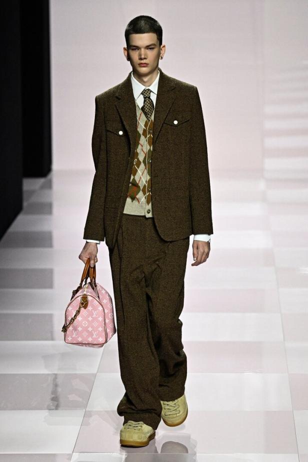 Ein Mann in einem braunen Anzug mit Argyle-Weste und einer rosa Louis Vuitton Tasche auf einem Laufsteg.