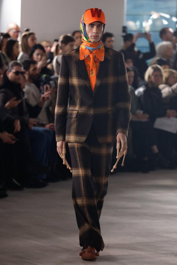 Ein Model in einem karierten Anzug mit orangefarbenem Hemd und Kappe auf dem Laufsteg.