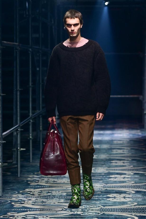 Ein Mann in einem schwarzen Pullover, braunen Hosen und gemusterten Stiefeln hält eine bordeauxrote Tasche.
