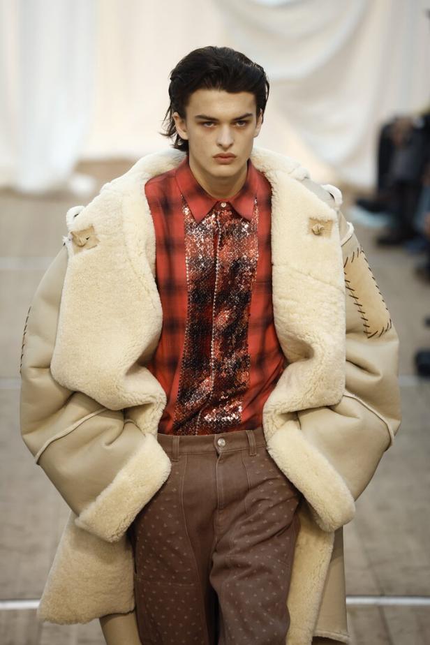 Ein Mann mit offenem Shearling-Mantel und rotem Hemd auf einem Laufsteg.